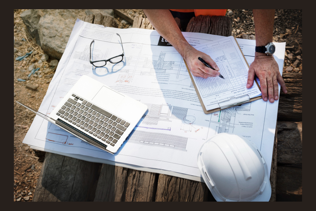 Contractor from a home improvement company in PA reviewing construction plans and filling out a checklist on-site in Mount Pleasant, PA, with a laptop, safety helmet, and architectural drawings.