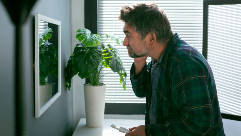 A man examining his face closely in a mirror in a modern room with natural light and plants, illustrating personal care and home improvement services.