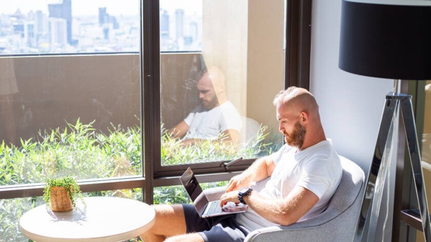 Man working on a laptop while sitting near large windows with natural light, showcasing the benefits of modern home improvement services for comfortable and bright living spaces.